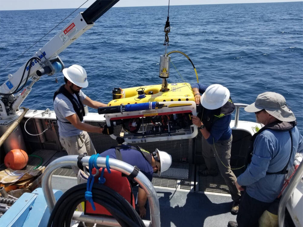 Four people on a boat handle a yellow autonomous underwater vehicle attached to a crane over open lake water.